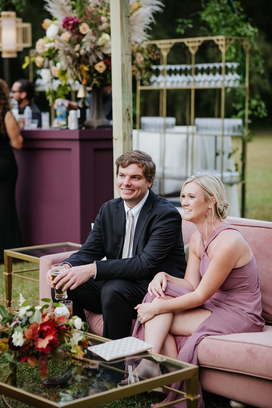 Couple seated on a pink couch at an outdoor event, smiling beside floral arrangements and a bar.