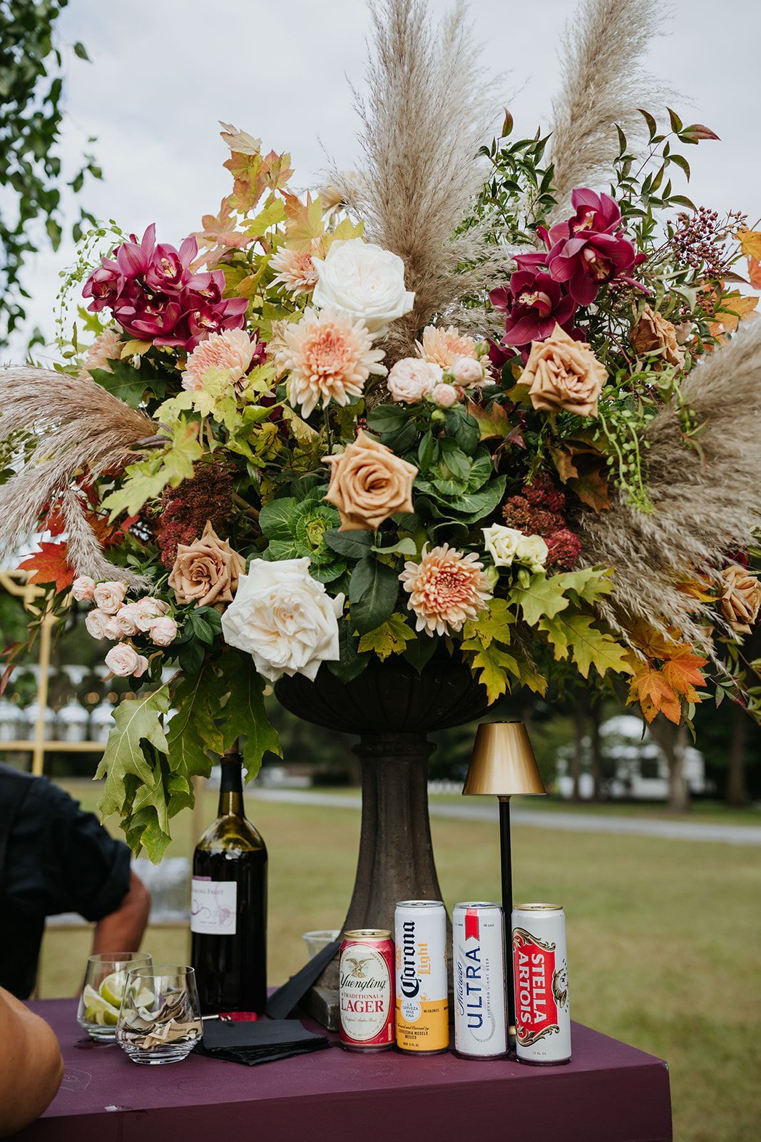 Floral centerpiece on a table with wine bottle, glasses, and cans at an outdoor event