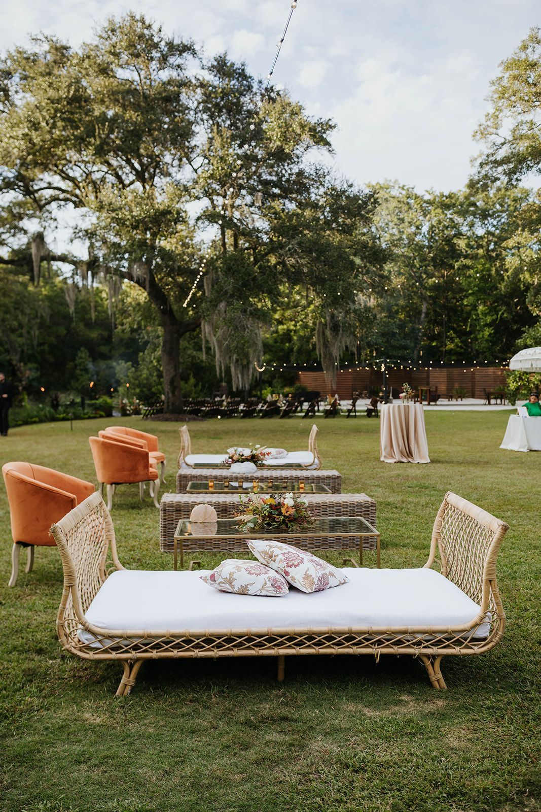 Outdoor lawn lounge with woven sofas, low tables, and orange cushions under trees at sunset