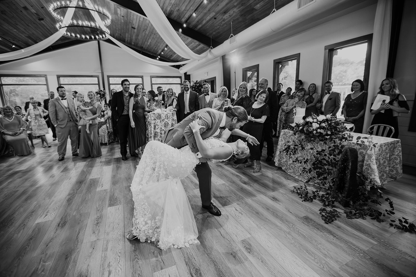 Couple dancing at a wedding reception in a decorated hall, surrounded by guests watching.