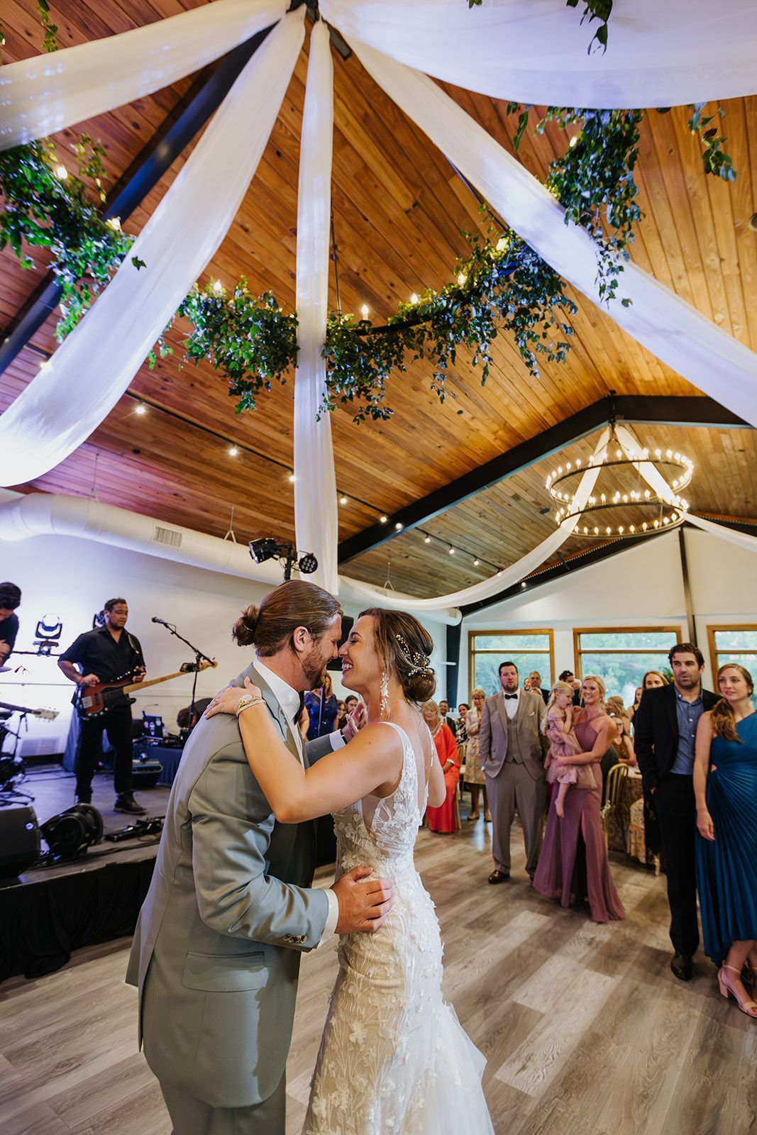 Bride and groom slow dance at a wedding reception under draped white fabric and warm string lights.
