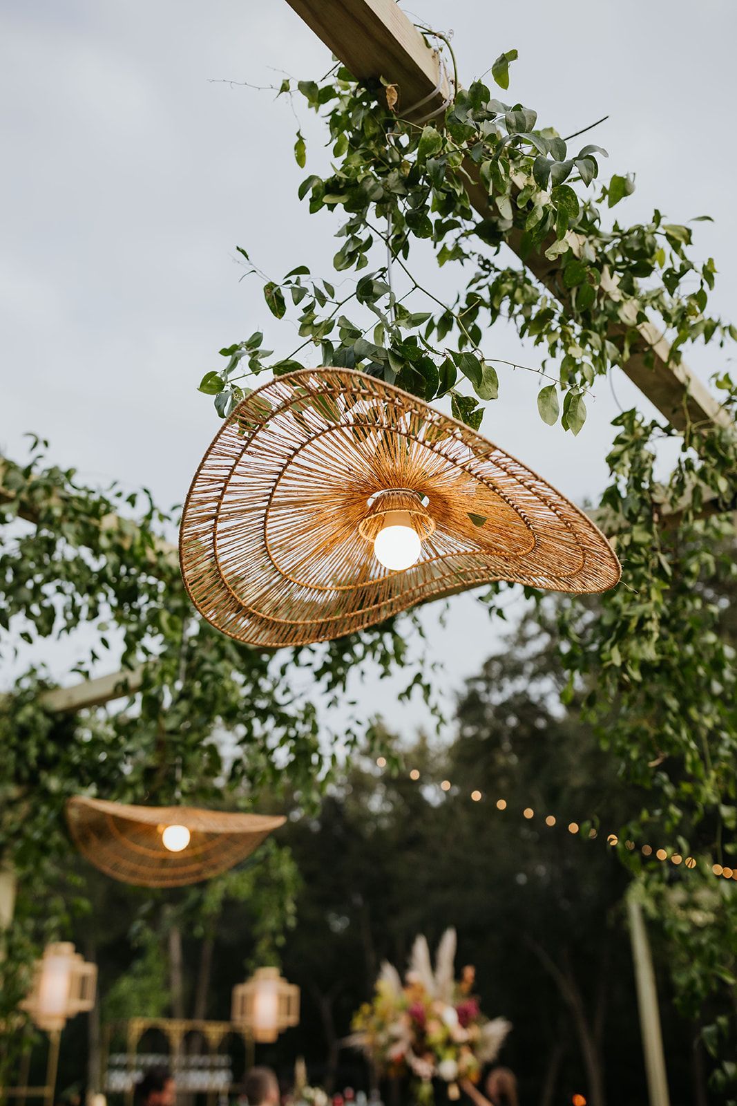 Woven pendant lamps hang from tree branches at an outdoor event.