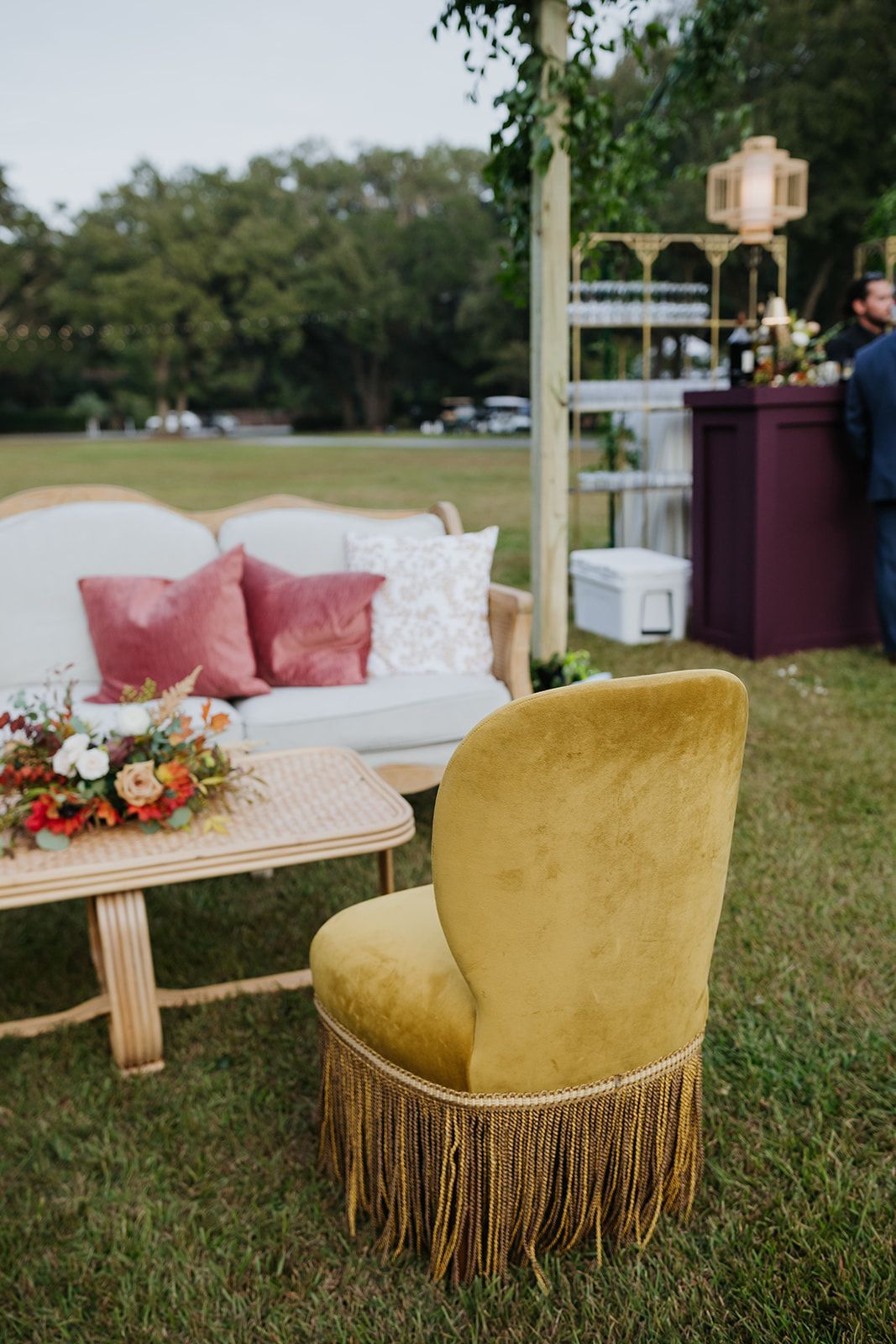 Outdoor lounge setup with gold fringed chair, white sofa, pink pillows, and floral table by a lake