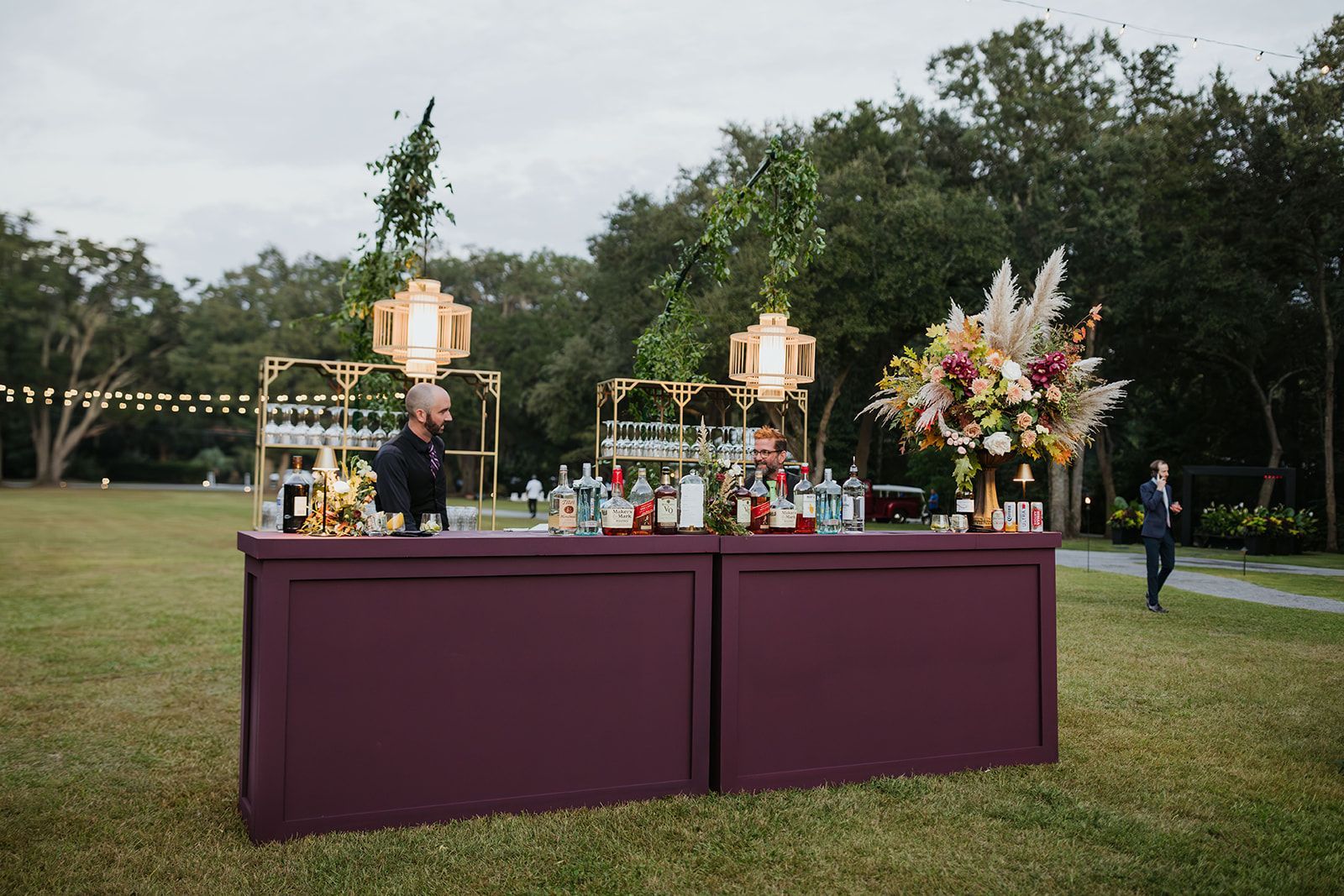 Outdoor bar setup with purple tablecloth, drinks, lanterns, and floral arrangements on a lawn at dusk