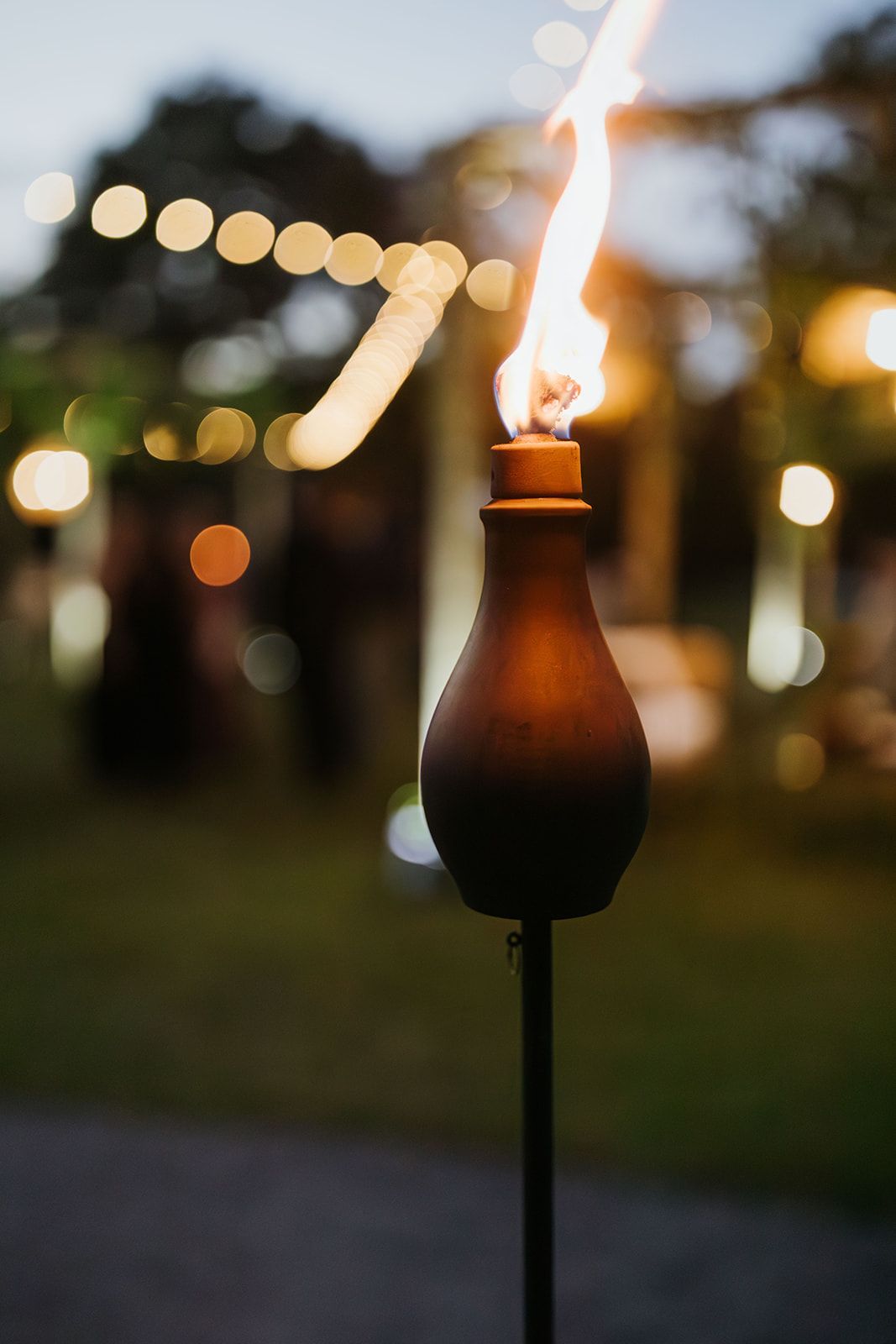 Lit tiki torch glowing at an outdoor evening event with blurred string lights in the background