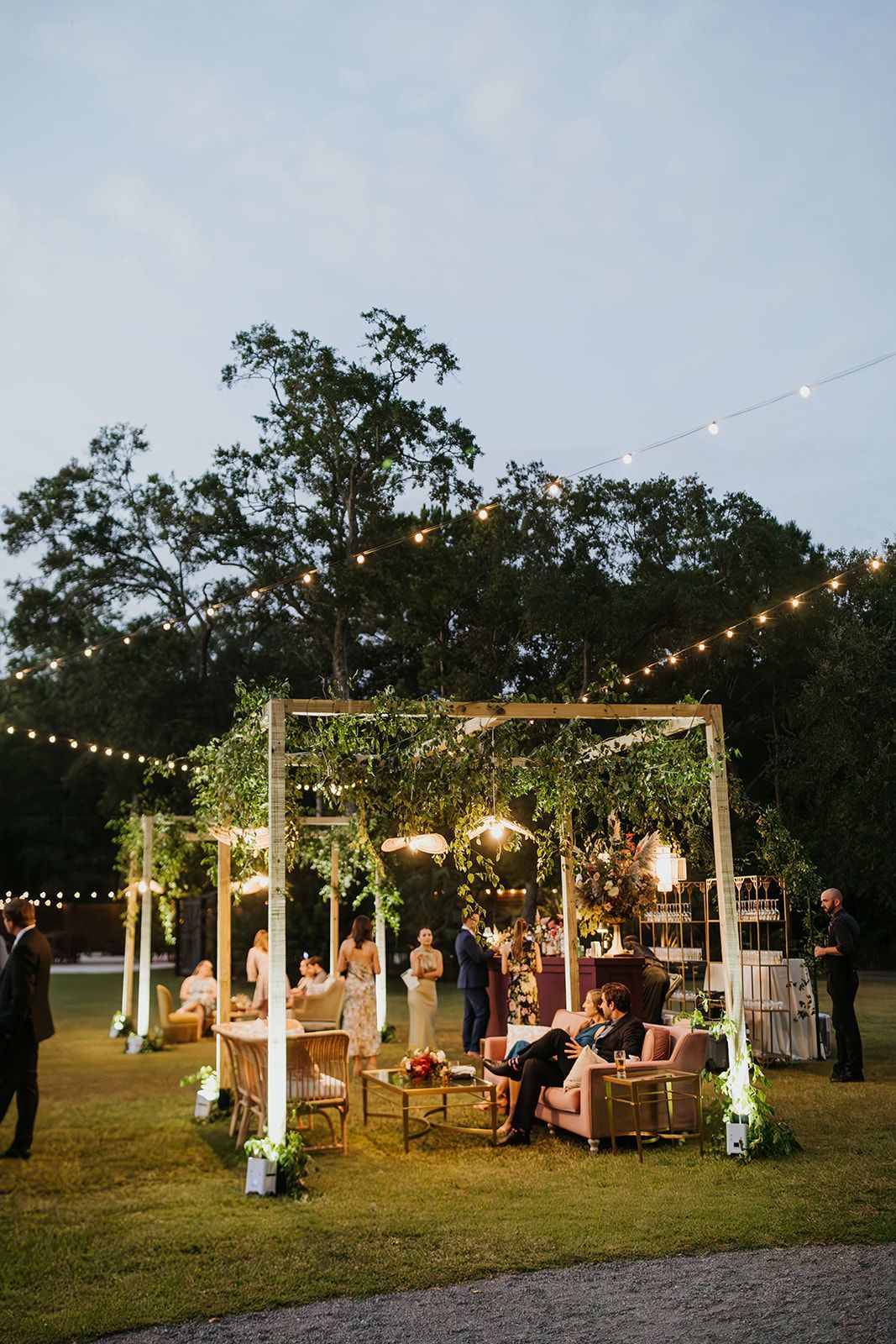 Outdoor evening gathering under string lights in a garden, with seated guests around a decorated wooden frame.