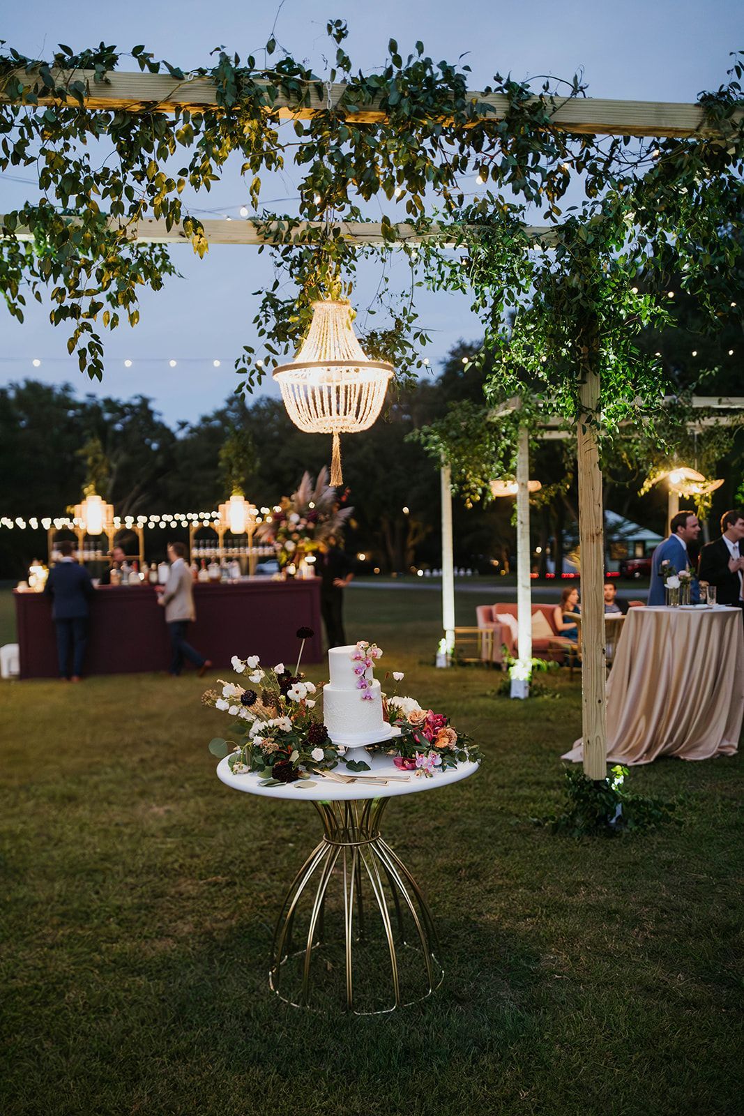 Outdoor evening reception with floral decorations, chandeliers, and guests at cocktail tables under a vine-covered arbor
