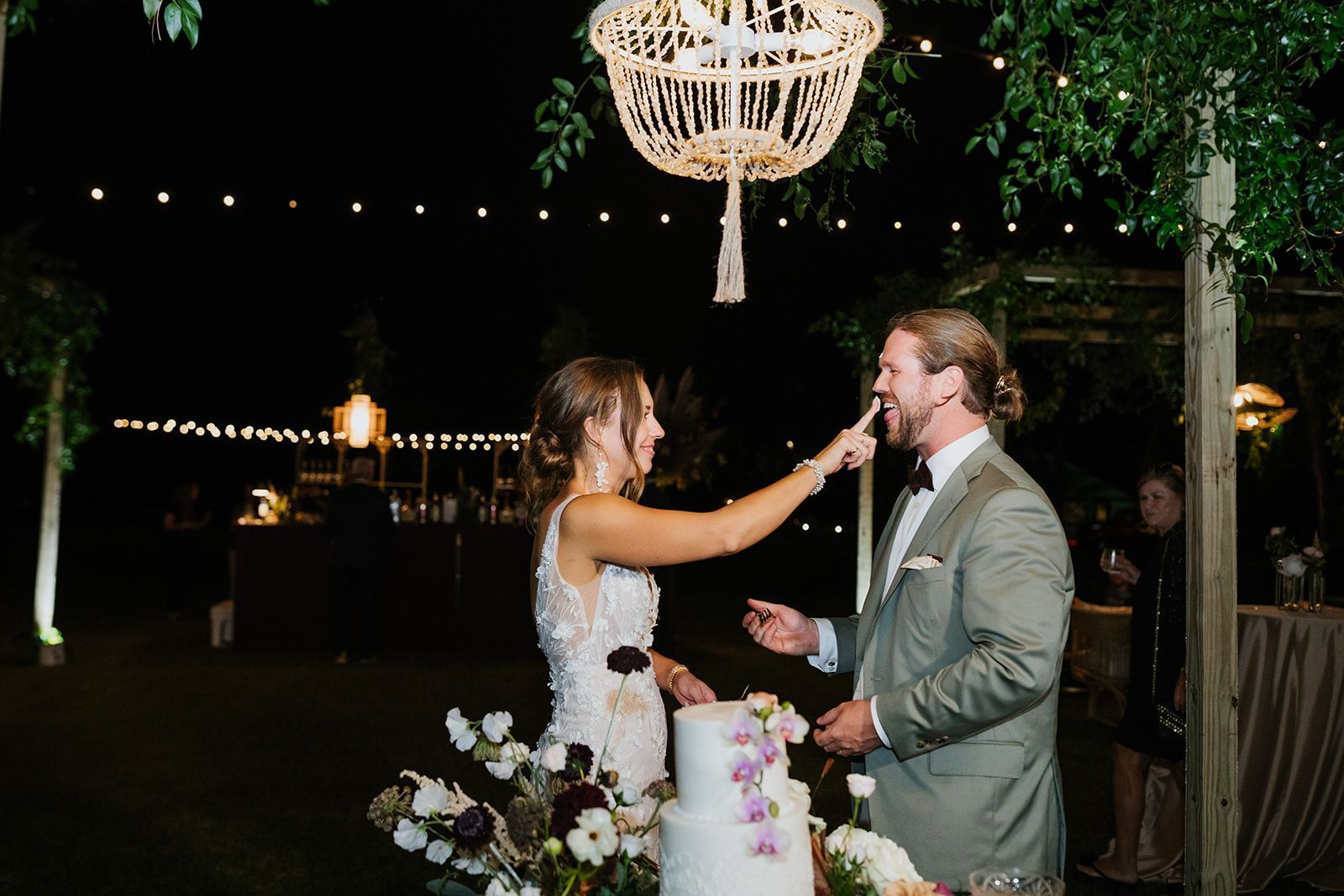 Bride feeding groom cake at an outdoor wedding reception under string lights