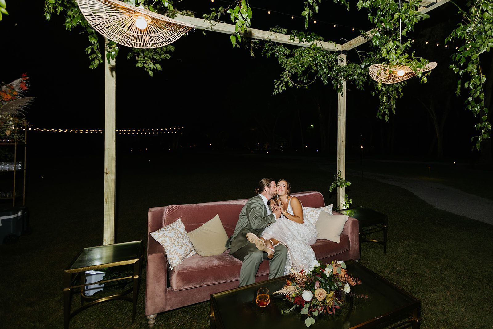 Couple on a pink sofa under a flowered canopy at night, with candles and floral arrangements nearby