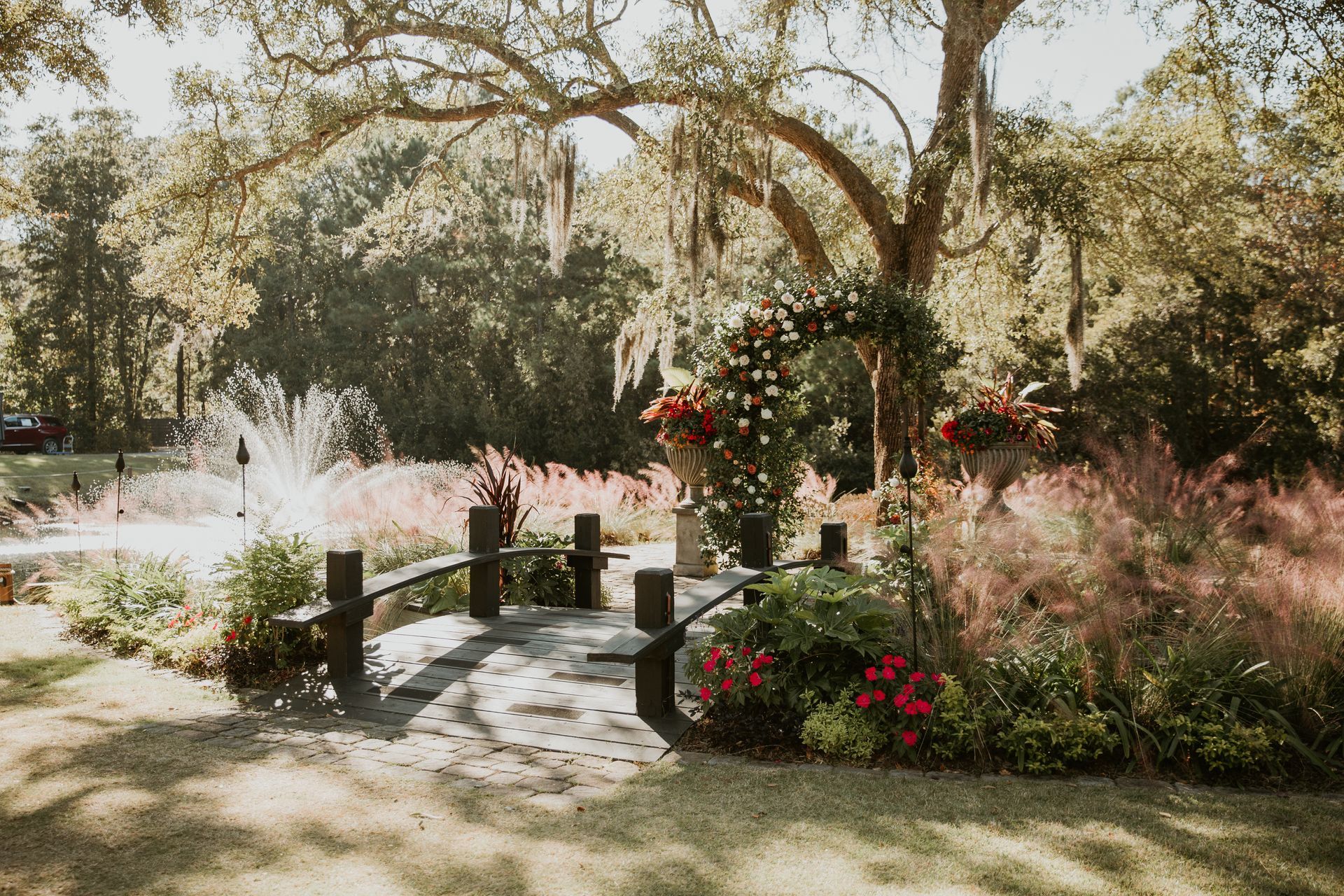 Sunny garden path with a small wooden bridge, trees, and flowering shrubs.
