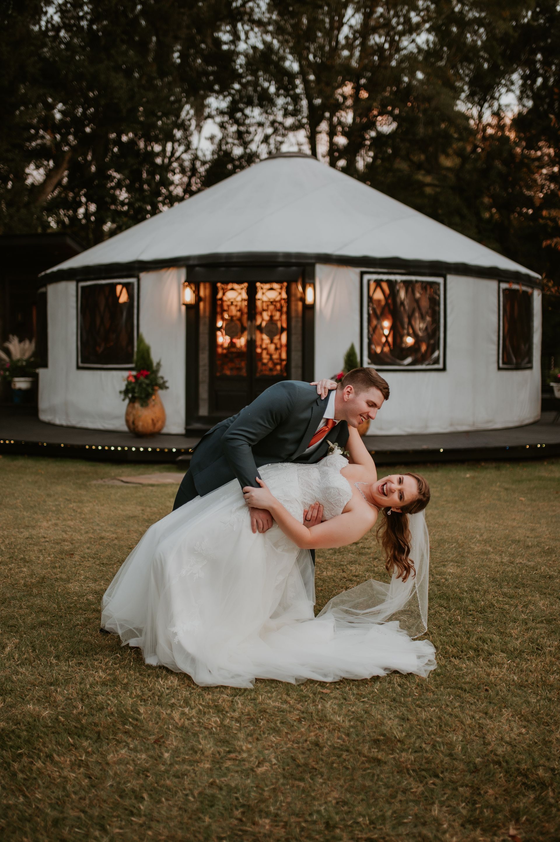 Couple in wedding attire posing in a dip on grass in front of a white round tent at dusk