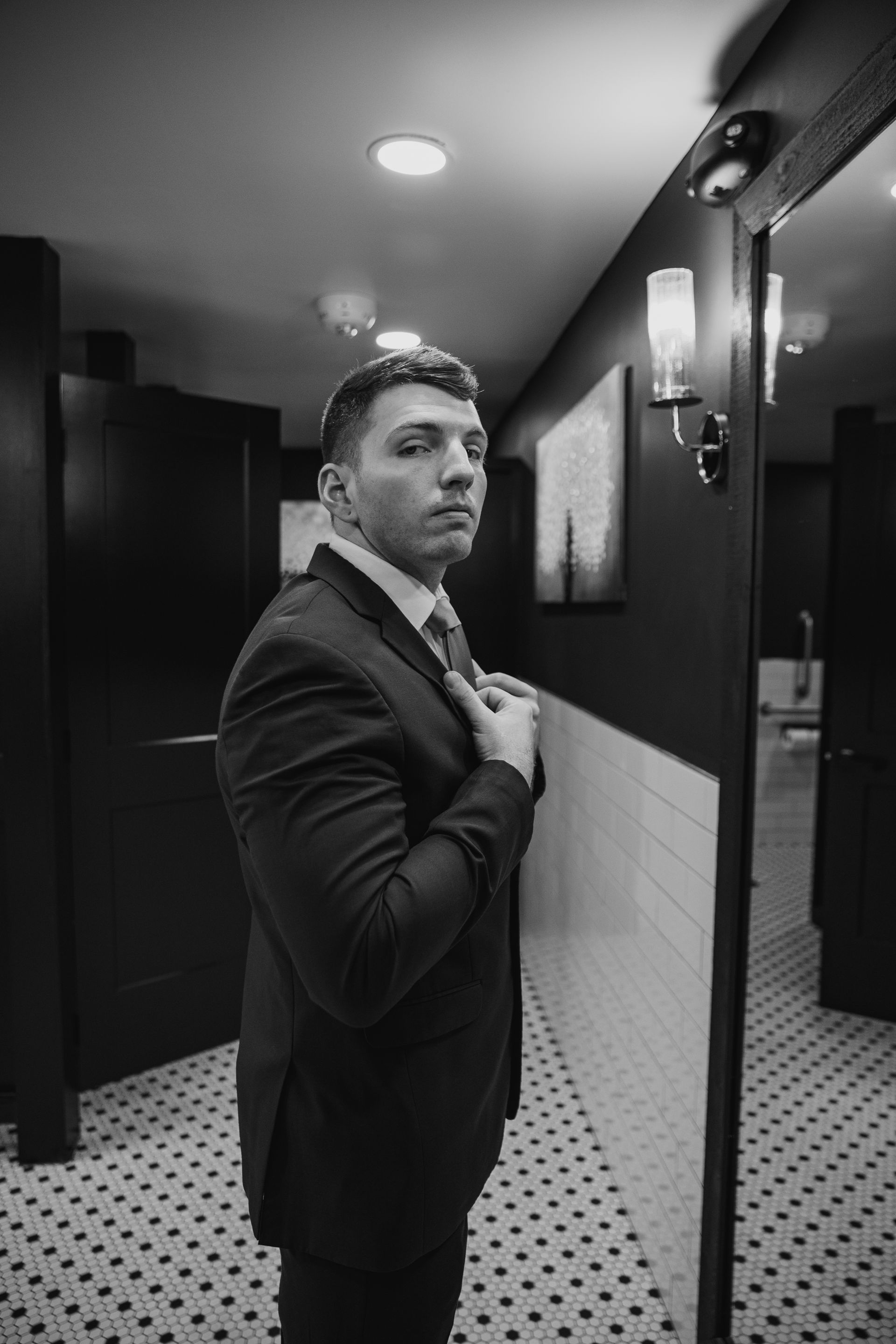 Man in a suit adjusting his tie in a hallway mirror, with patterned floor and wall lights.