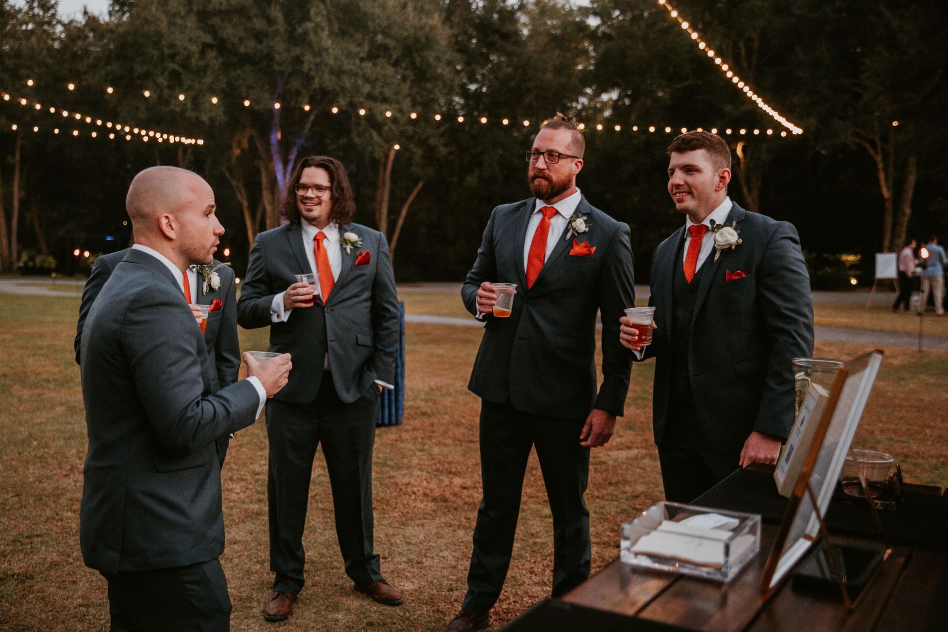 Four men in suits holding drinks at an outdoor evening gathering with string lights.