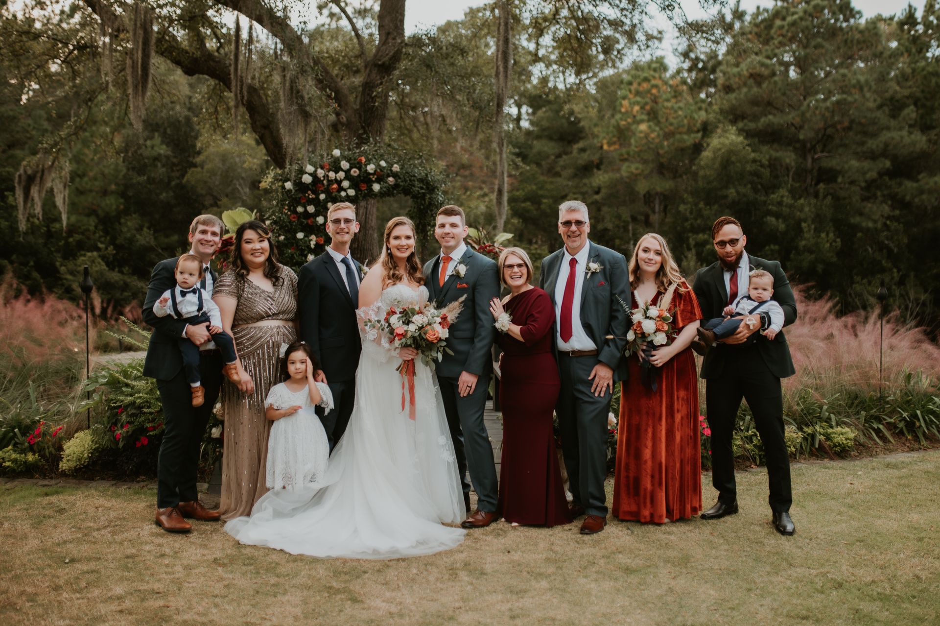 Wedding party posing outdoors in formal attire, bride holding bouquet under a floral arch.
