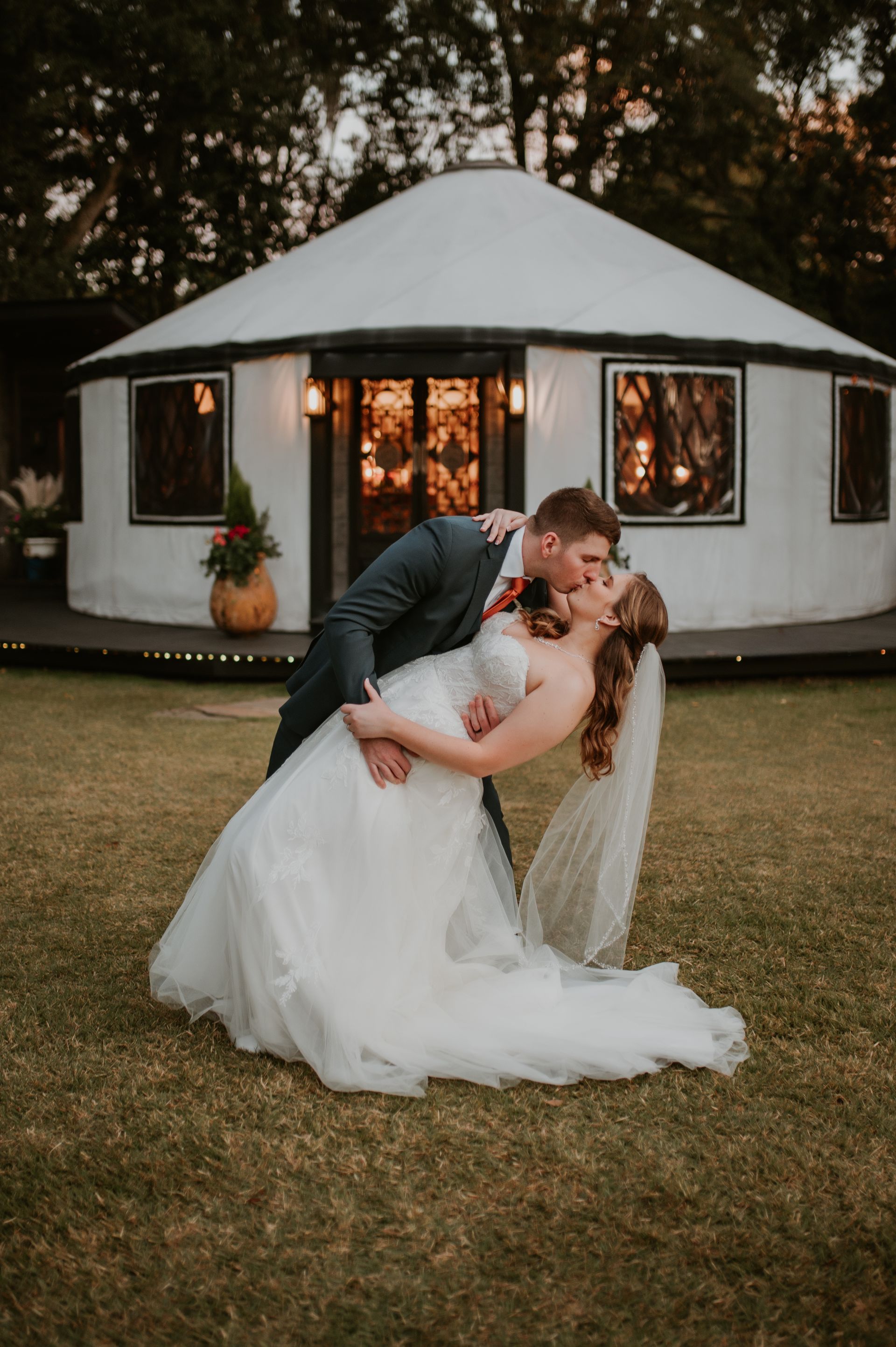Bride and groom kissing on grass in front of a lit white pavilion at dusk