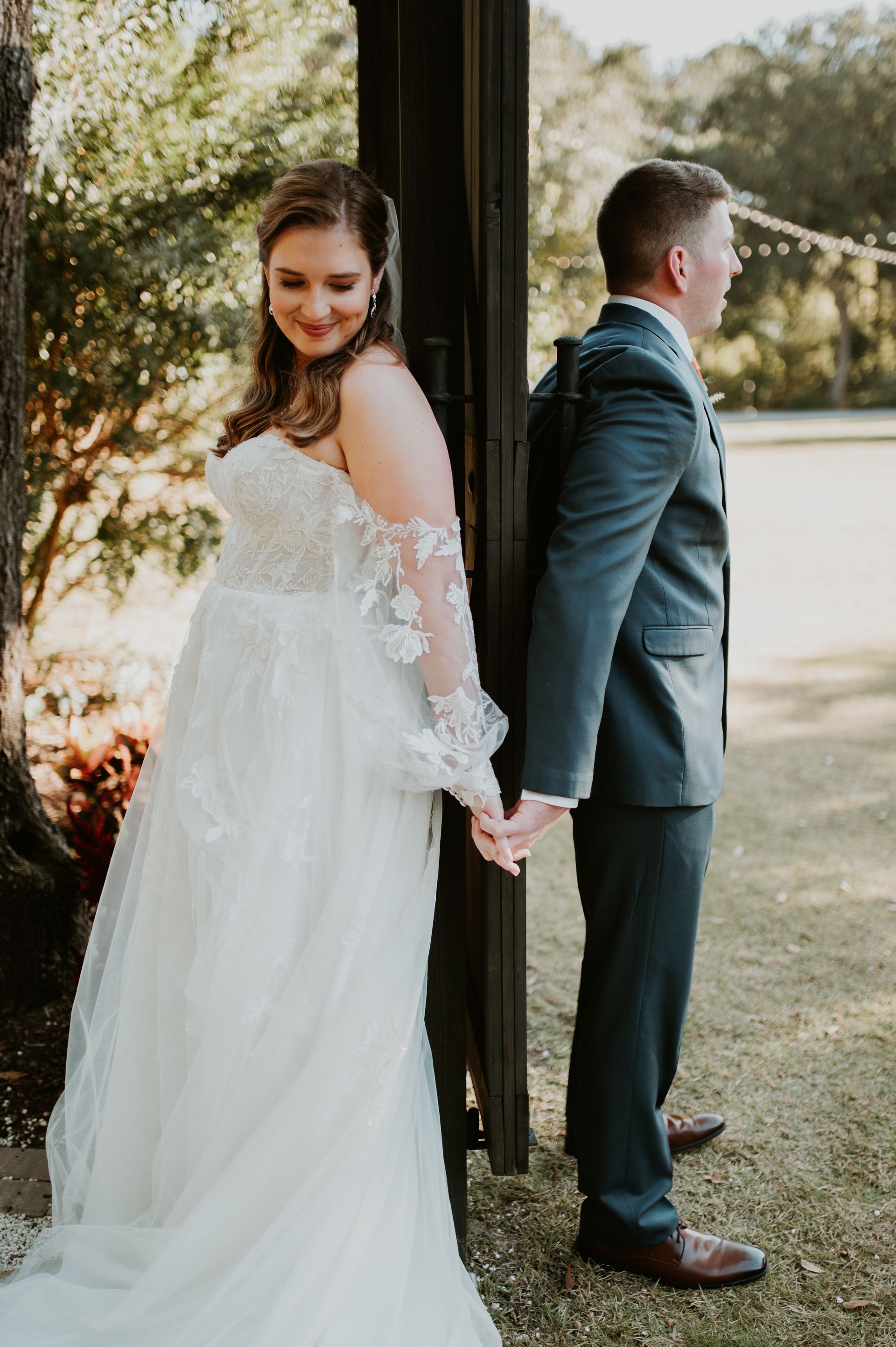 Bride in white gown and groom in blue suit holding hands back-to-back by a tree outdoors