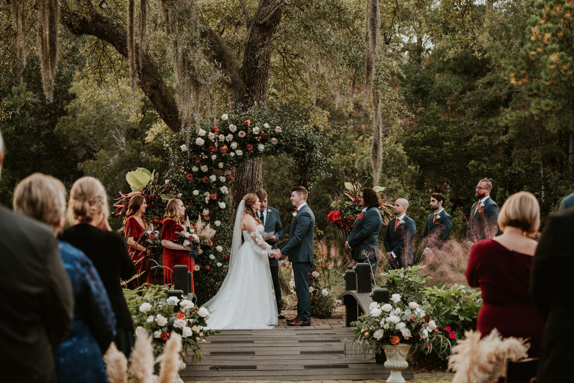 Outdoor wedding ceremony with bride and groom under a floral arch, surrounded by guests and greenery.