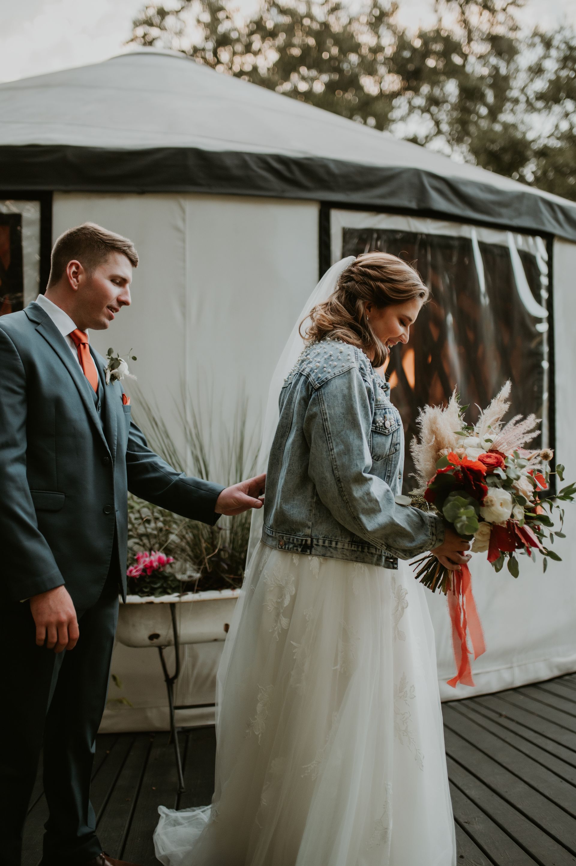 Bride and groom walking outdoors beside a white tent, holding a bouquet and wearing formal wedding attire