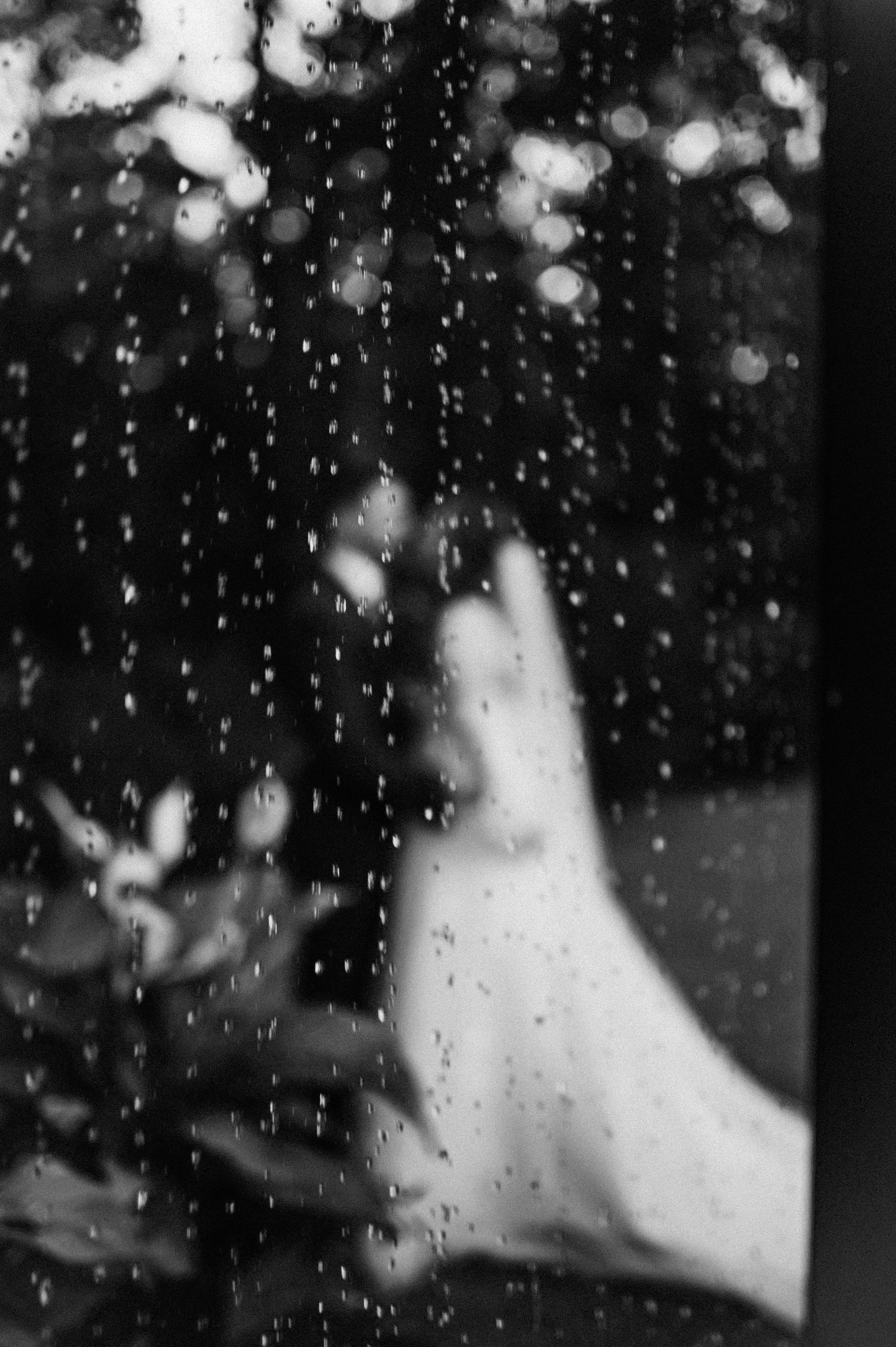 A foot pressing against a rain-speckled window beside leafy plants in black and white.