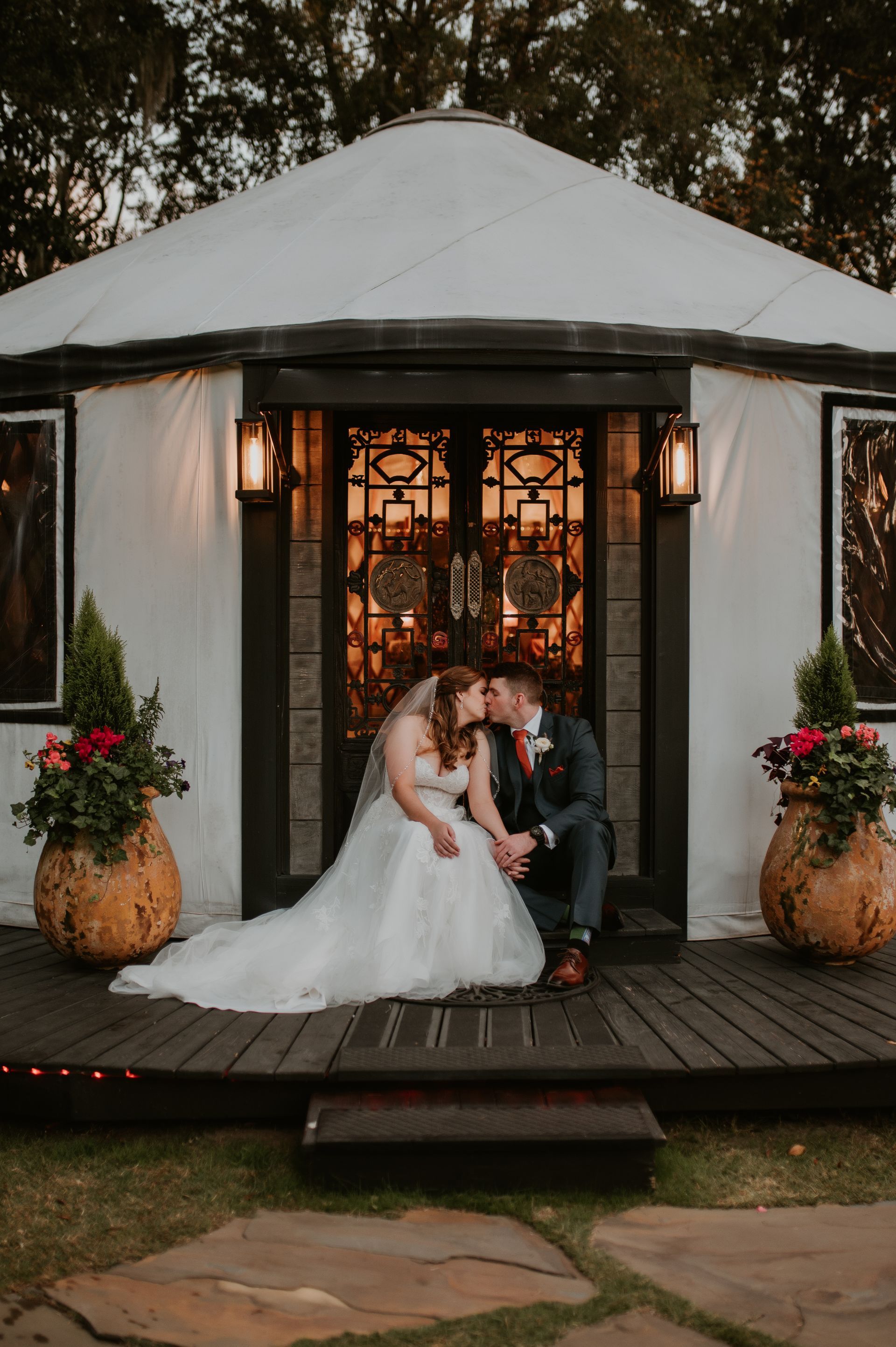 Newlyweds seated together on a lit gazebo porch with floral arrangements at dusk