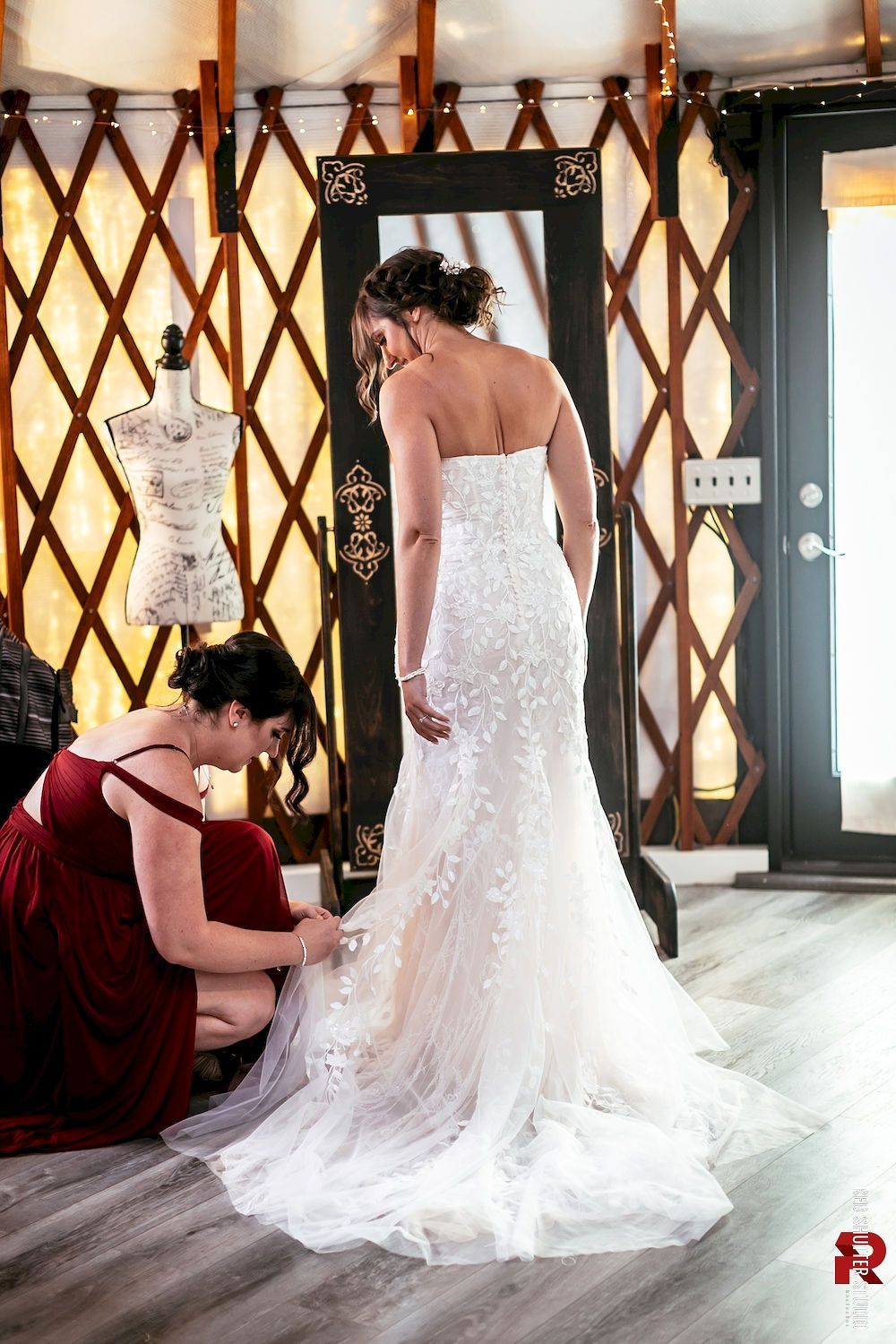 Bride in a lace wedding gown being helped by a woman in a burgundy dress in a patterned room