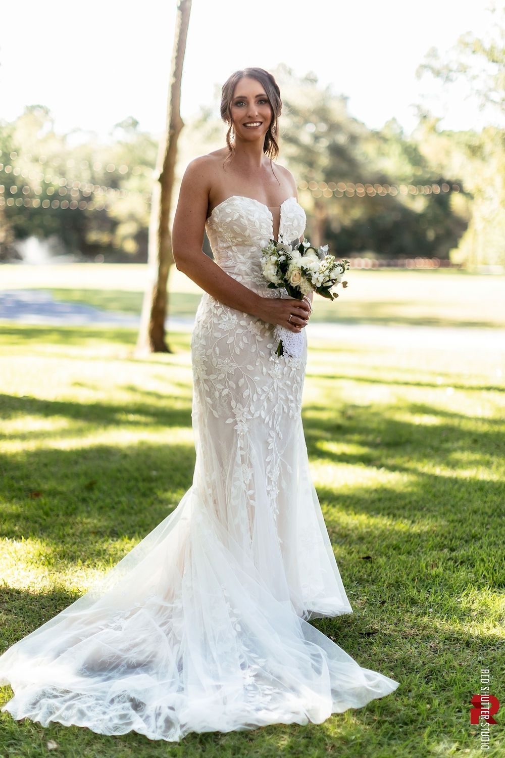 Bride in a white lace wedding gown holding a bouquet outdoors in a sunlit park