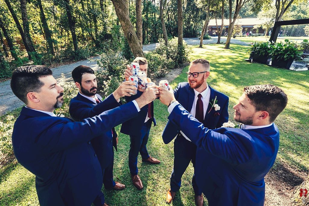 Five men in suits toast with glasses in a sunny park beneath trees.