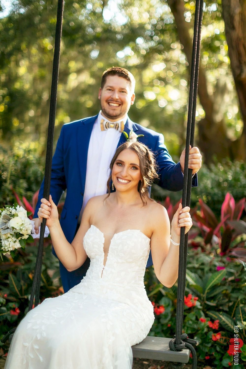 Bride and groom smiling on a swing in a garden, with greenery and flowers behind them