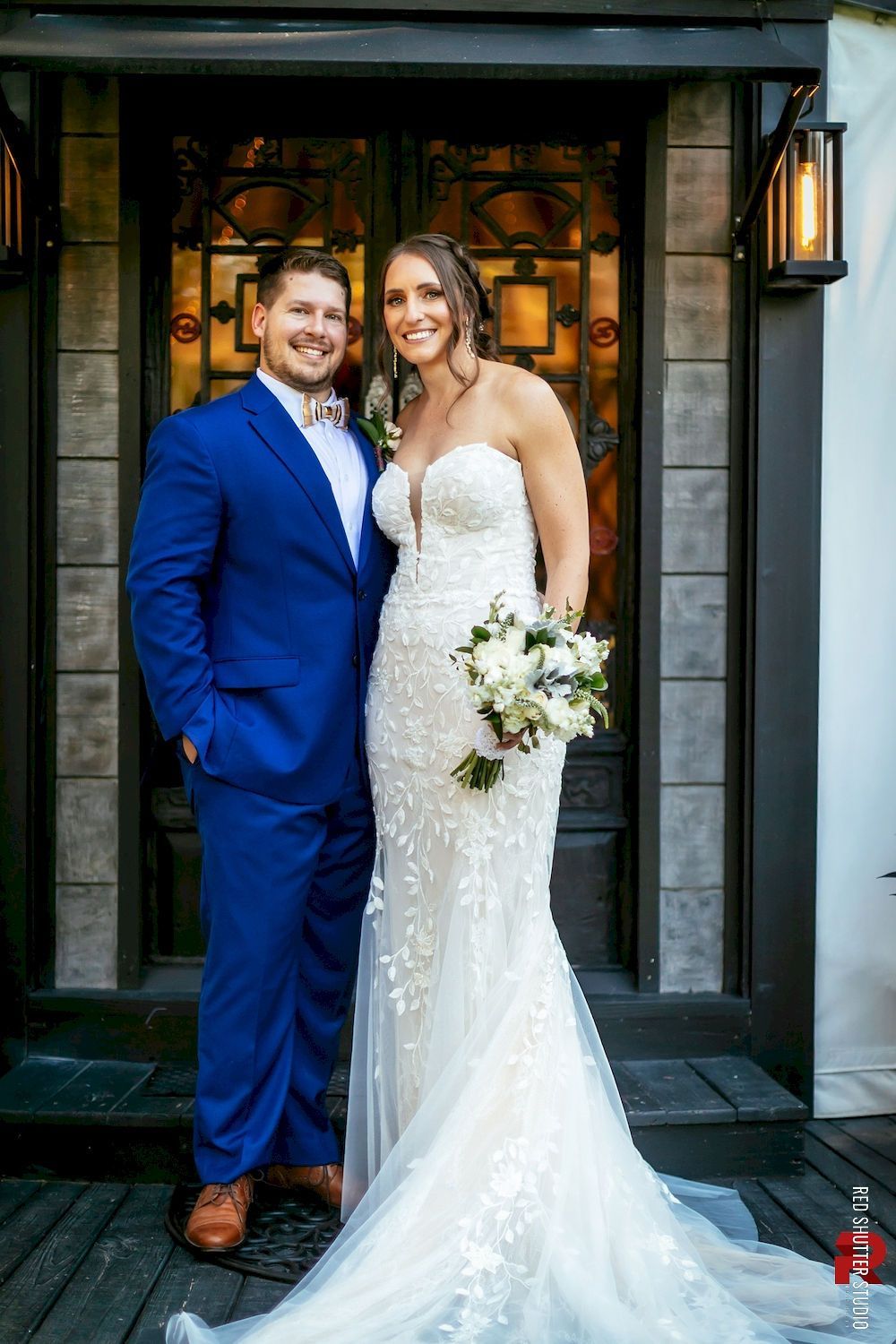 Bride and groom posing outside a doorway, with the bride in a white lace gown and the groom in a blue suit.