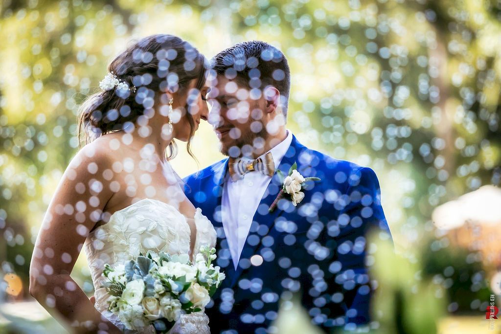 Wedding couple embracing under falling confetti, bride holding white bouquet, groom in blue suit outdoors