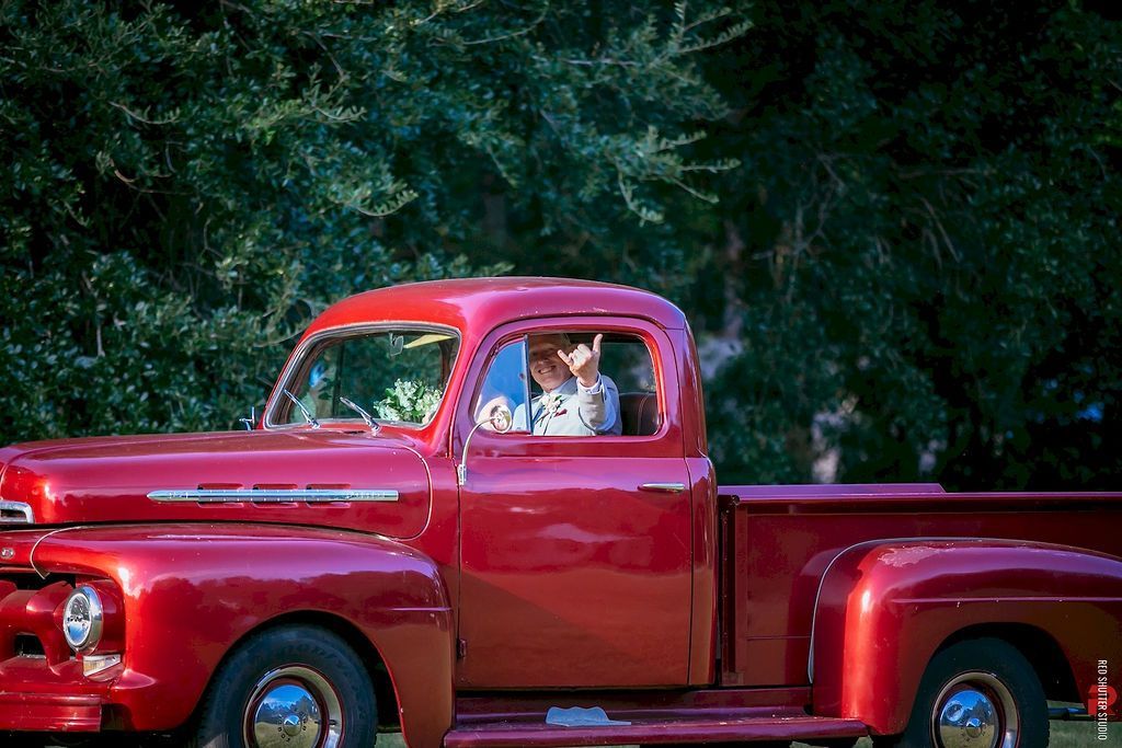 Red vintage pickup truck with two people in the cab, parked against a leafy green background
