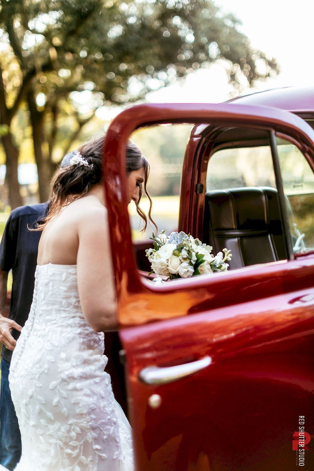 Bride in a white gown leaning into a red vintage car with a bouquet nearby outside