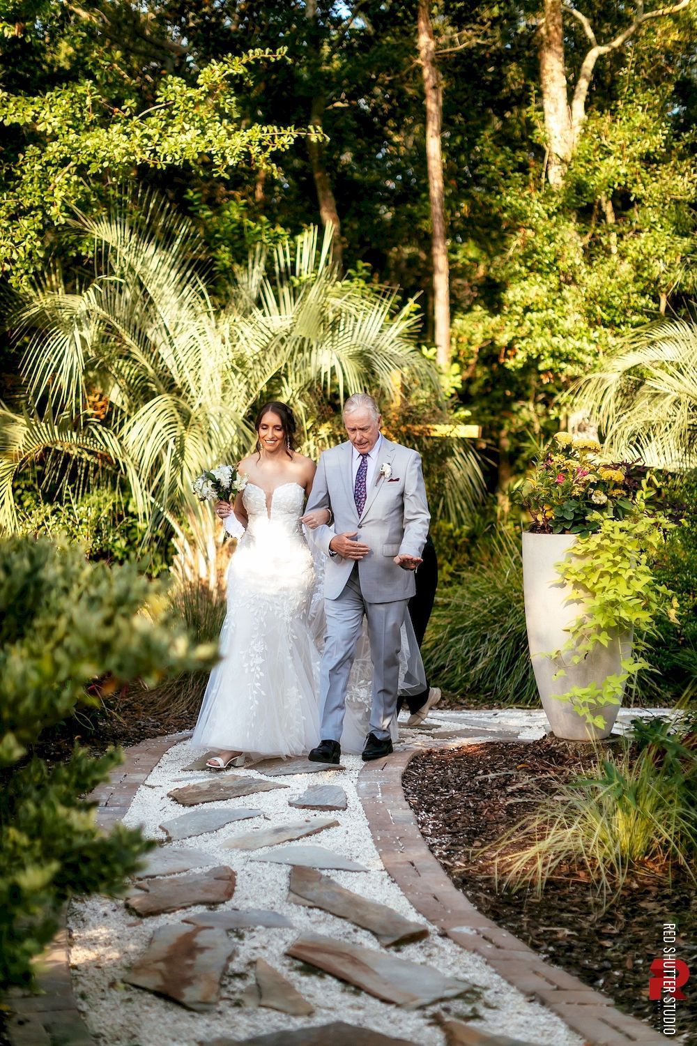 Newlyweds walking down a stone garden path surrounded by lush greenery, holding hands and smiling