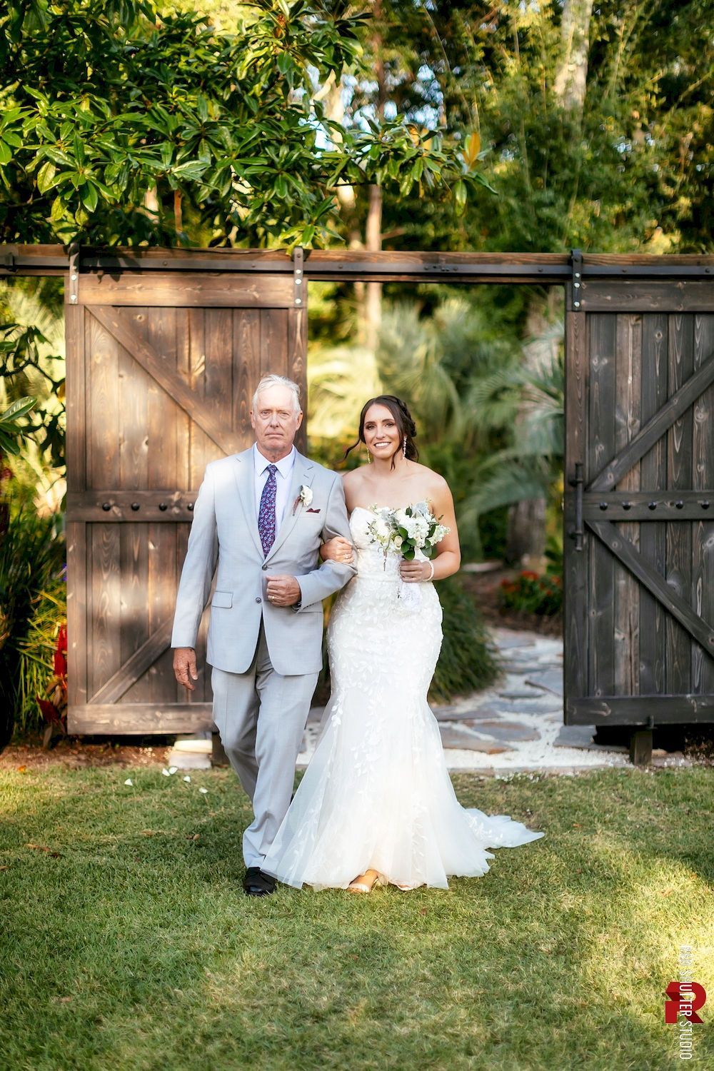 Bride and groom walk through wooden gates on a sunlit lawn, holding flowers and smiling.