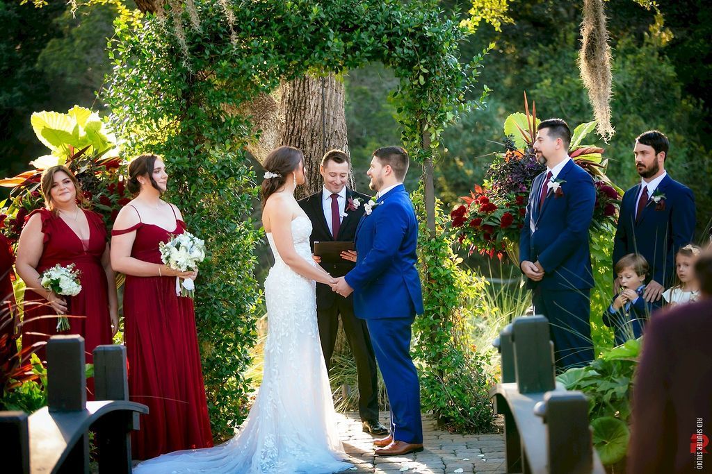 Wedding ceremony outdoors under a leafy arch, bride in white and groom in blue exchanging vows, attendants nearby