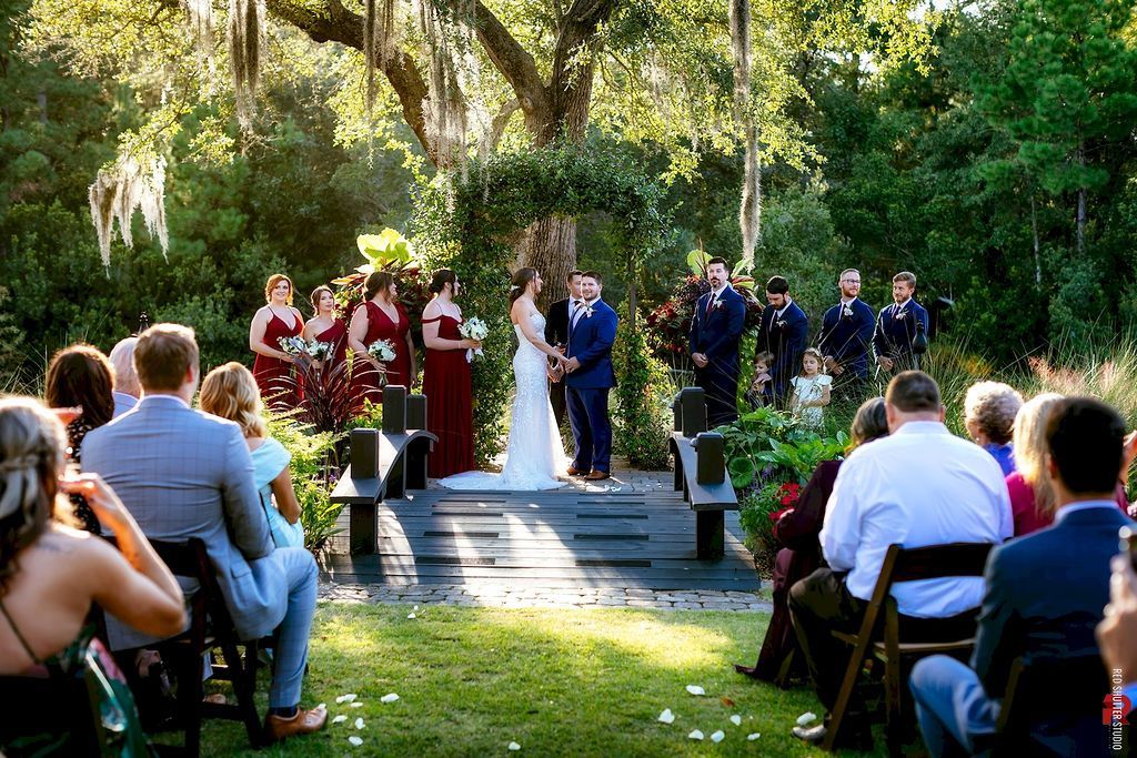Outdoor wedding ceremony with bride and groom under trees, guests seated on either side of the aisle.