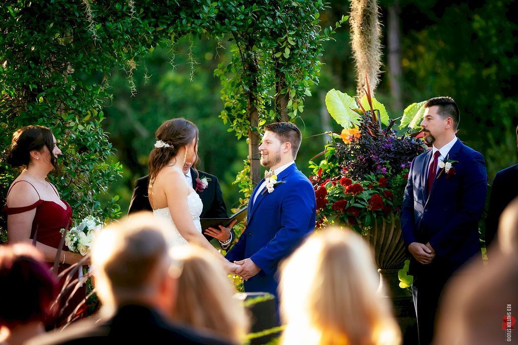 Bride and groom exchange vows outdoors beside a colorful floral arrangement under green trees