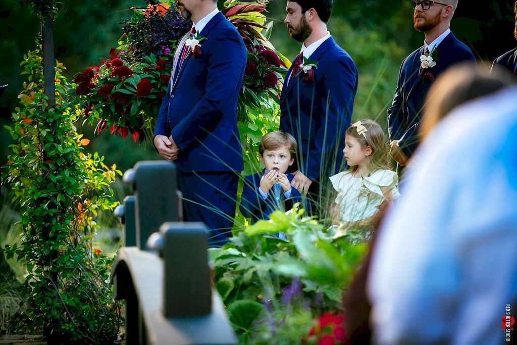 People in blue suits stand by two children in a flower-filled garden aisle.