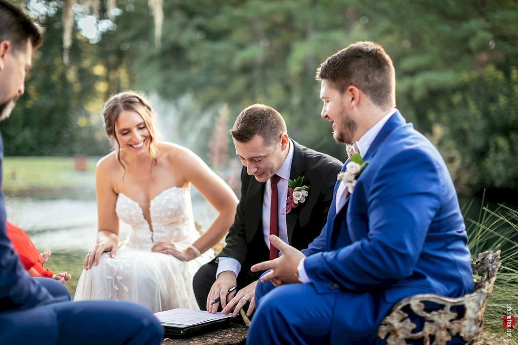 Wedding party seated outdoors by a small fire, with the bride in a white dress and a groom in a blue suit.