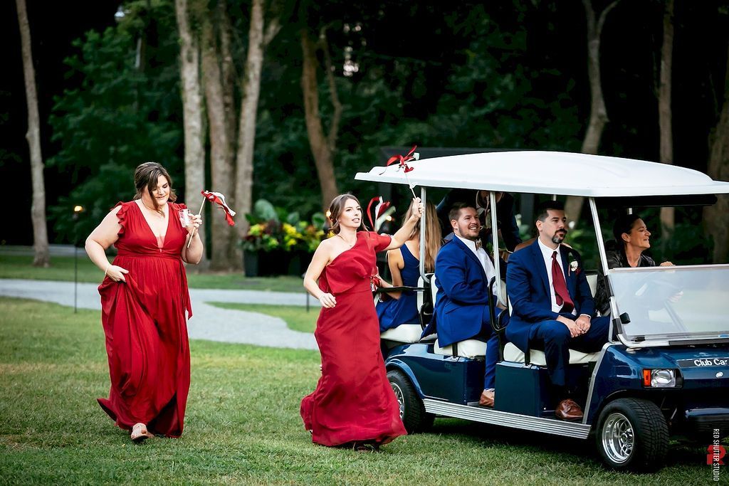 Three people in red dresses escort men in blue suits on a golf cart outdoors on a grassy path.