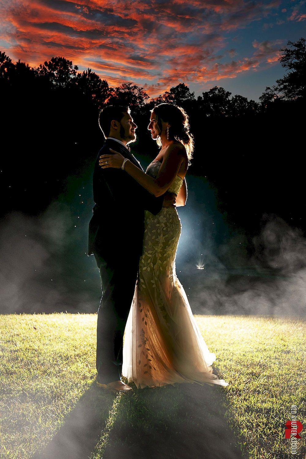 Backlit couple embracing in a misty field at sunset under a dramatic red sky