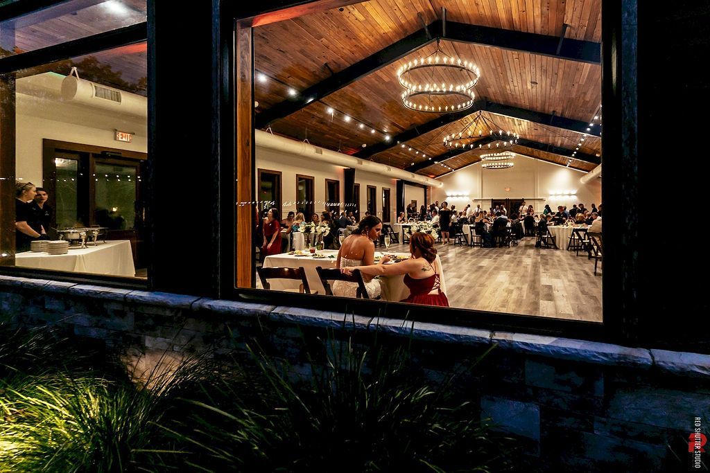 A modern event hall with a glowing chandelier, seated guests, and a polished wooden floor seen through a large window