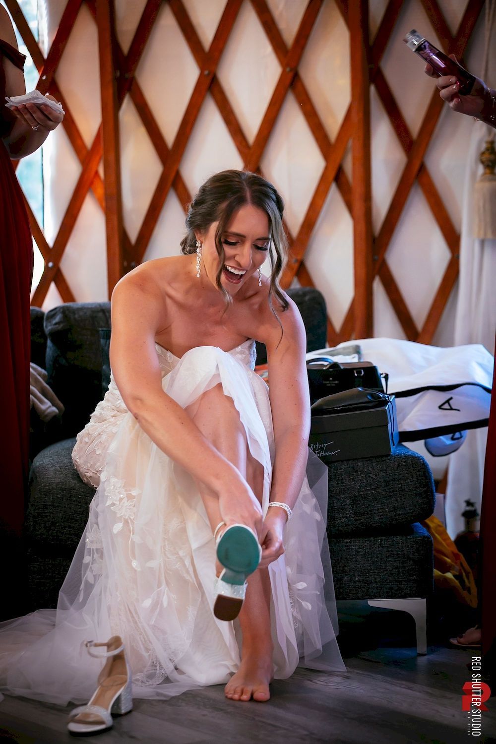 Bride putting on a white shoe while seated in a bridal gown indoors.