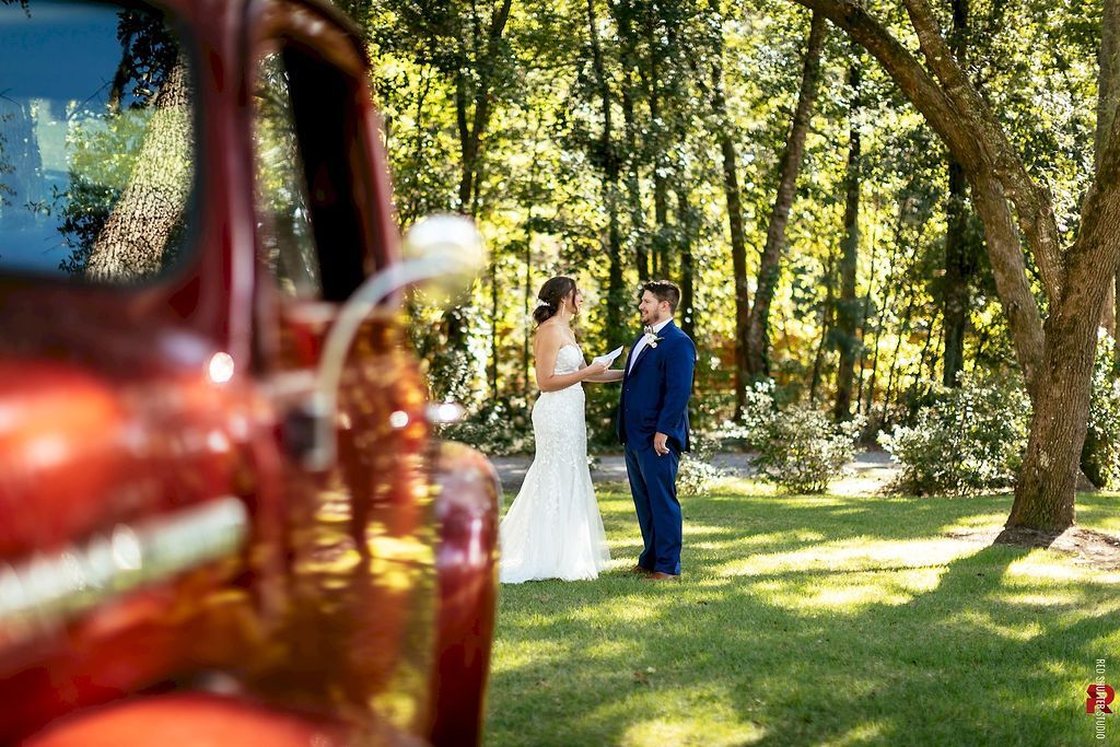 Bride and groom standing in a sunlit garden, with a red car blurred in the foreground