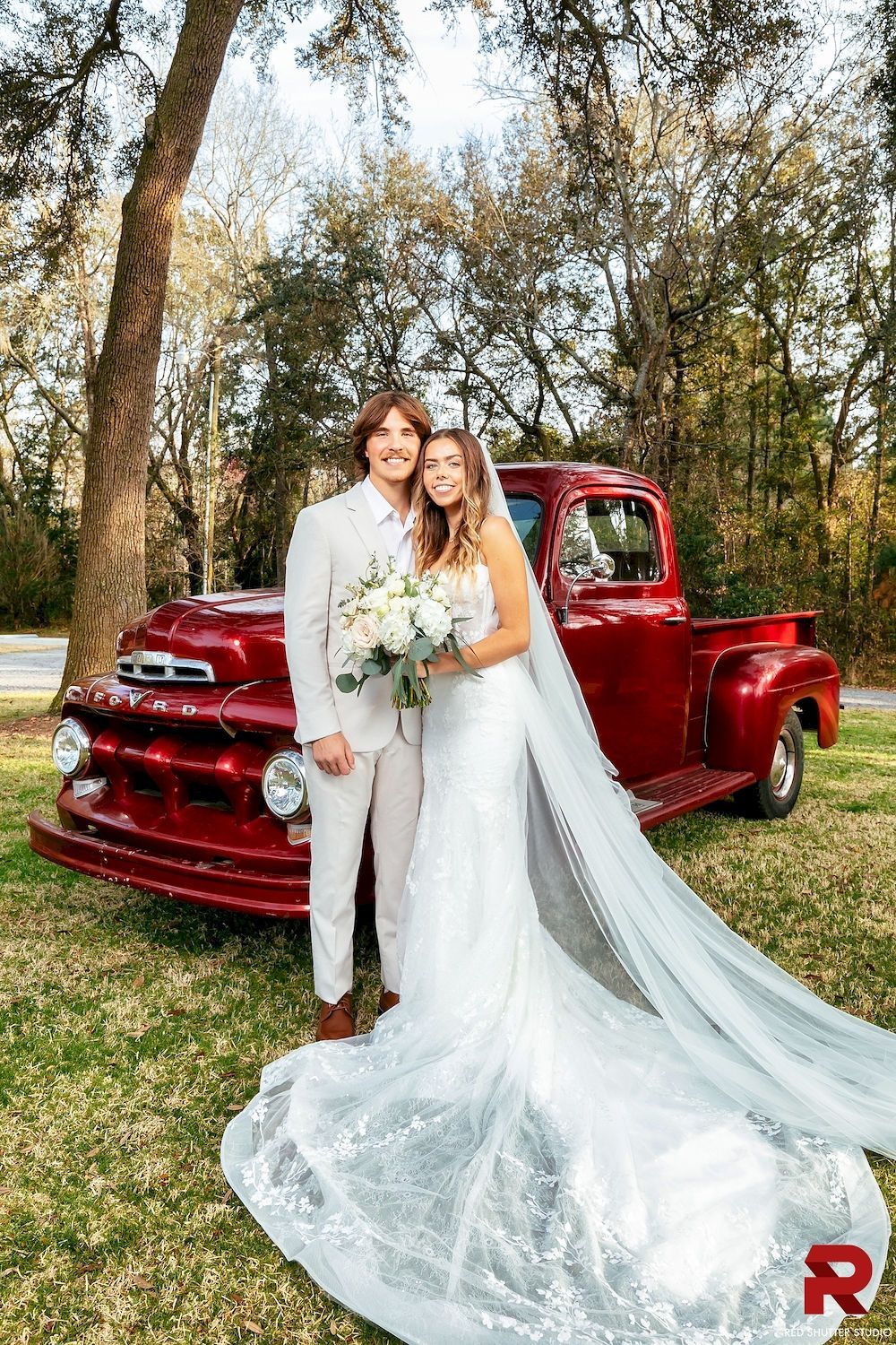 Bride and groom posing beside a vintage red truck in a wooded outdoor setting