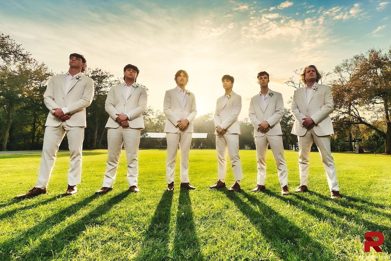 Six people in white suits stand on grass at sunset, with long shadows and a bright sky behind them.