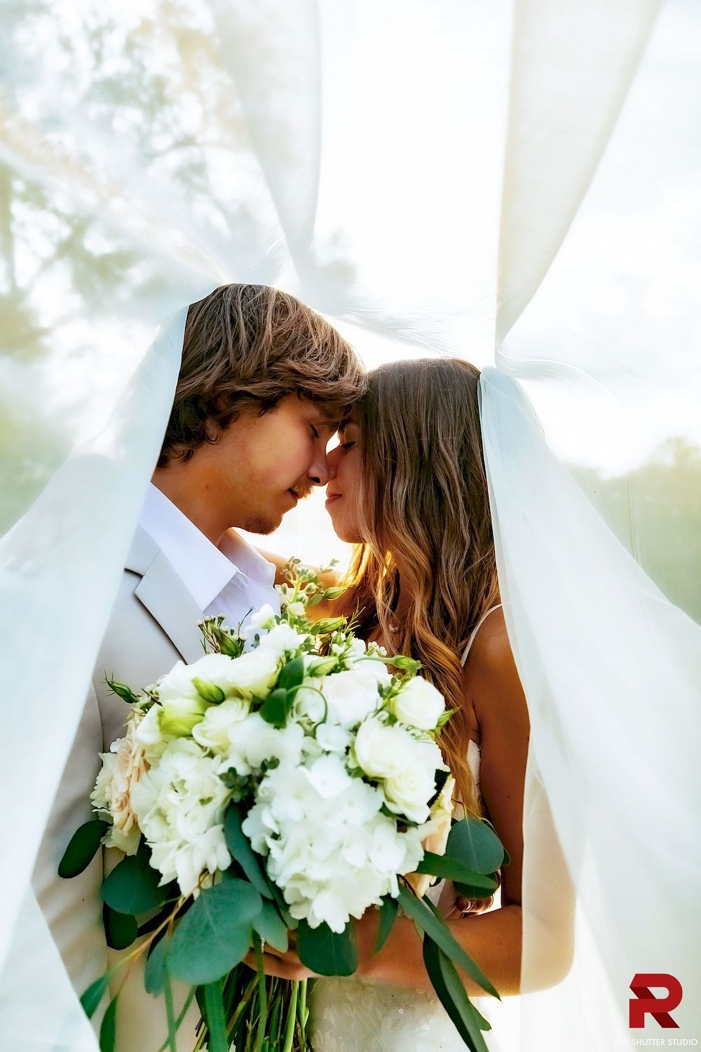 Couple embracing under white drapes, holding a bouquet of white flowers.