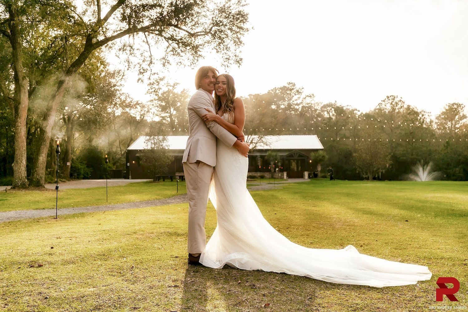 Couple in wedding attire embracing outdoors on a grassy lawn at sunset.