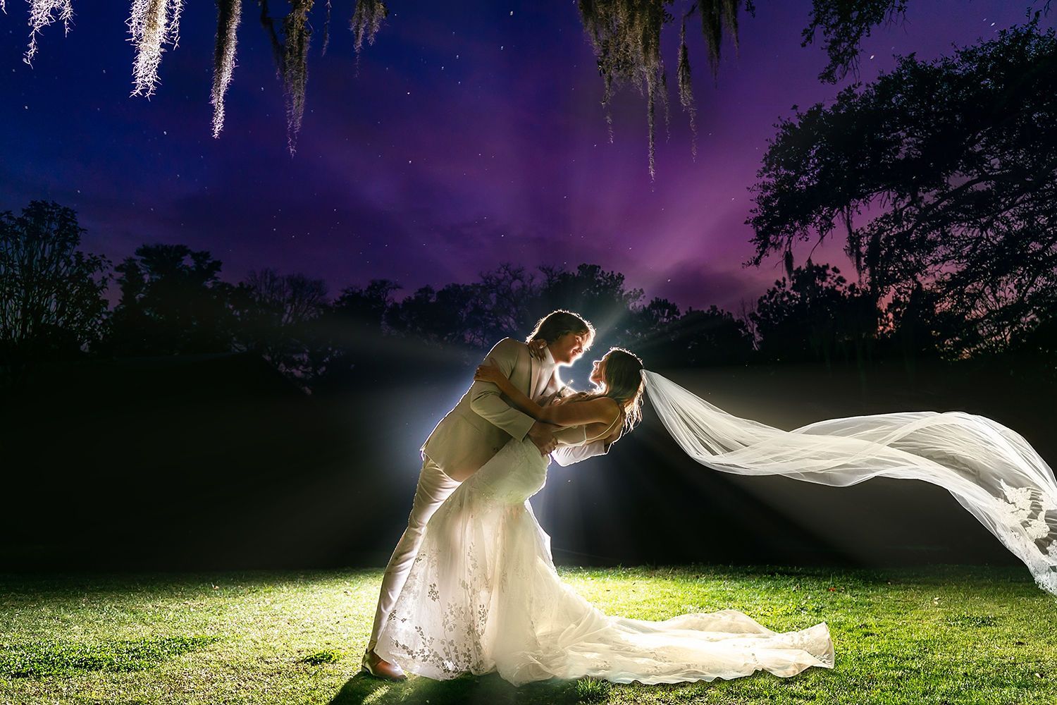 Bride and groom embrace in a moonlit garden, her white veil flowing under purple fireworks sky.