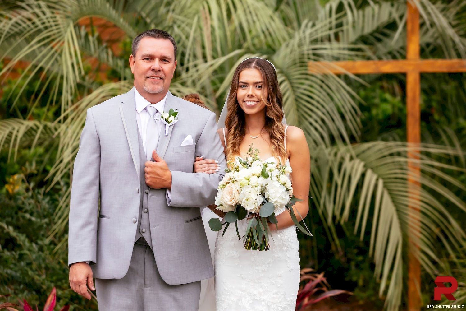 Bride and groom posing outdoors with bouquet before lush greenery and a wooden cross backdrop